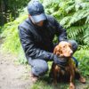 A man in a dark jacket and cap crouching on a woodland path beside a reddish-brown spaniel with damp fur, holding the dog's blue lead as ferns fill the background.