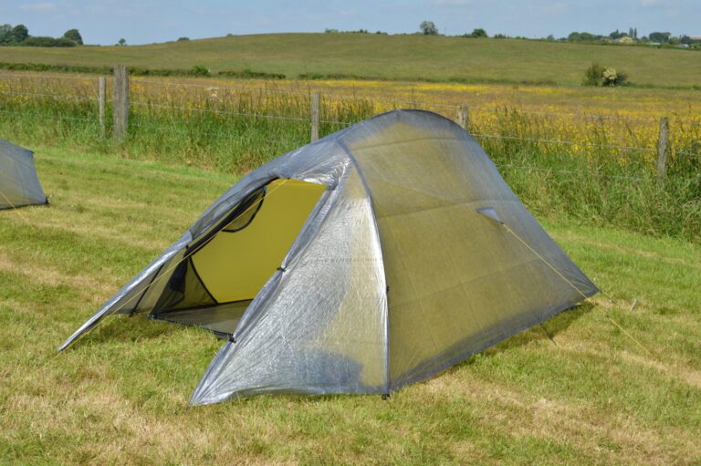 Lightweight backpacking tent with a translucent grey fly and yellow inner pitched on a grassy field beside a wire fence, with rolling hills and yellow wildflowers in the background.