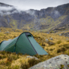 A green backpacking tent pitched on grassy, rocky moorland with steep, mist-shrouded mountains and low grey clouds in the background.