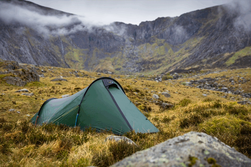 A green backpacking tent pitched on grassy, rocky moorland with steep, mist-shrouded mountains and low grey clouds in the background.