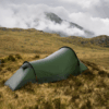 A lone olive-green tunnel tent pitched on windswept brown grass amid rocky moorland, with low cloud and mist rolling over distant mountains under an overcast sky.