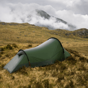 A lone olive-green tunnel tent pitched on windswept brown grass amid rocky moorland, with low cloud and mist rolling over distant mountains under an overcast sky.