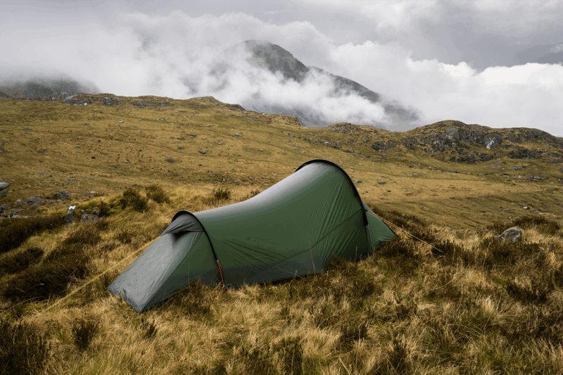 A lone olive-green tunnel tent pitched on windswept brown grass amid rocky moorland, with low cloud and mist rolling over distant mountains under an overcast sky.