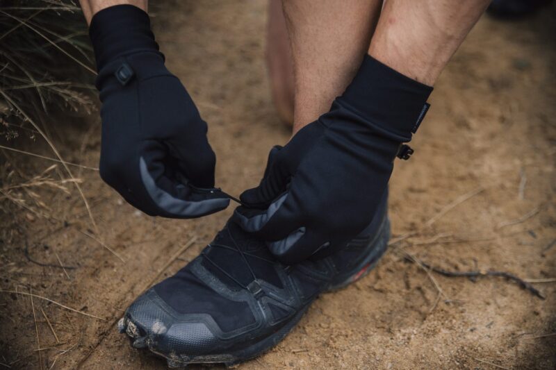 Gloved hands tightening the quick-lace on a scuffed black trail running shoe while standing on sandy ground.