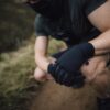 Crouching person in dark athletic clothing and a neck gaiter with gloved hands clasped over a knee on a dirt trail, showing muscular forearms and a blurred grassy background.