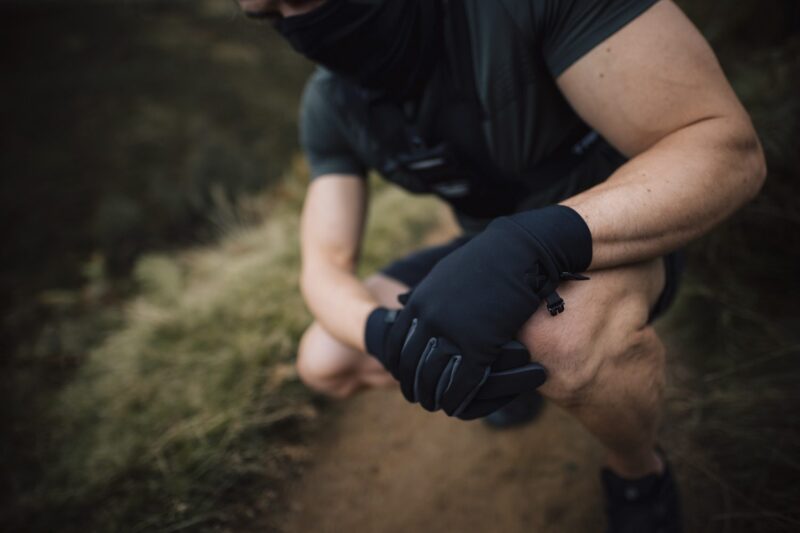 Crouching person in dark athletic clothing and a neck gaiter with gloved hands clasped over a knee on a dirt trail, showing muscular forearms and a blurred grassy background.