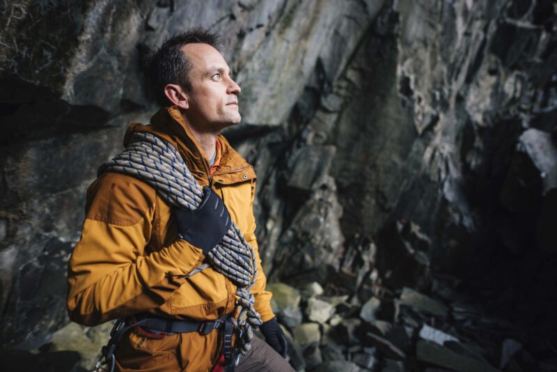 Man in an orange jacket and gloves stands inside a rocky cave holding coiled climbing rope over his shoulder, wearing a climbing harness and looking up at the cliff face.