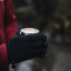 Person in a red fleece jacket wearing black gloves holding a small metal cup of creamy hot drink against a blurred outdoor background