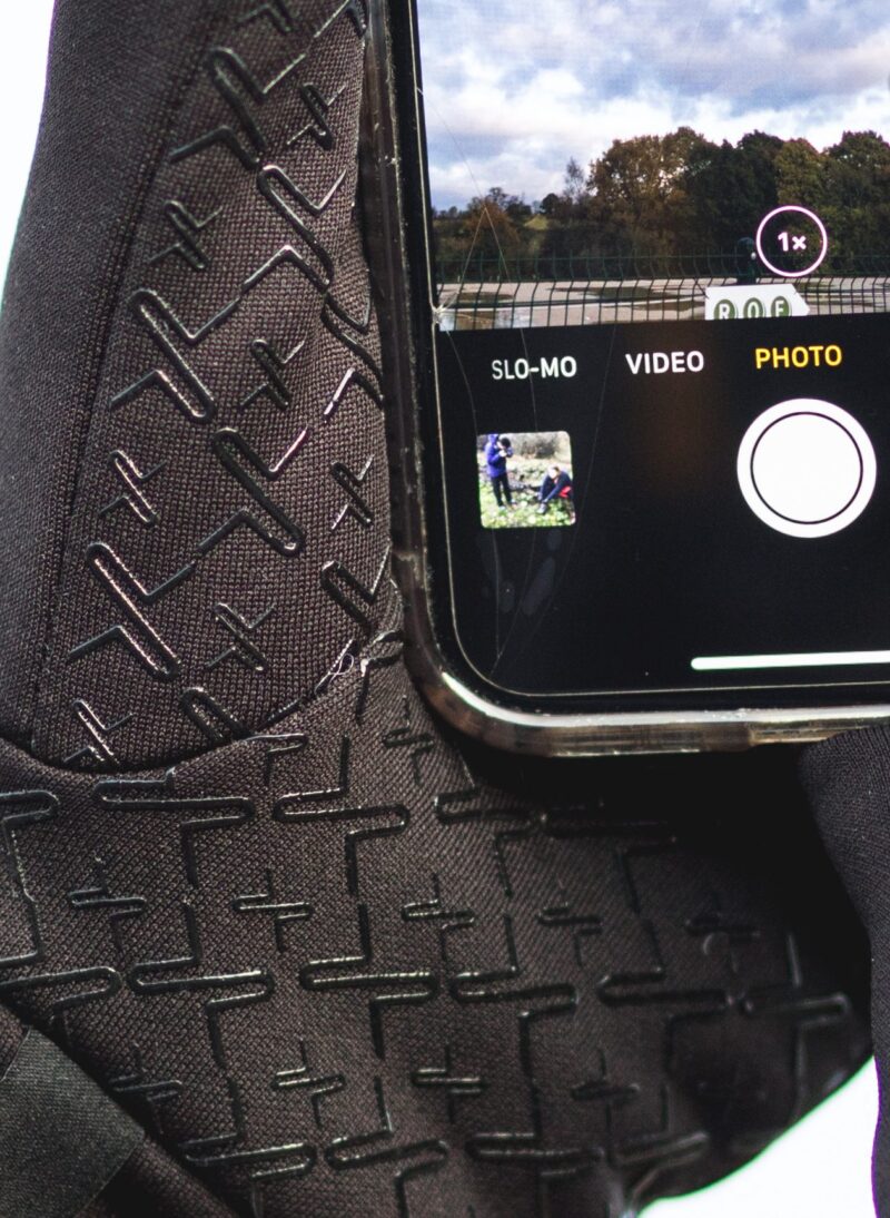Close-up of a smartphone resting on textured black fabric with a raised plus-shaped pattern, showing the camera app in Photo mode with a white circular shutter, 1x zoom indicator, a small landscape preview and a faint crack across the phone's glass.
