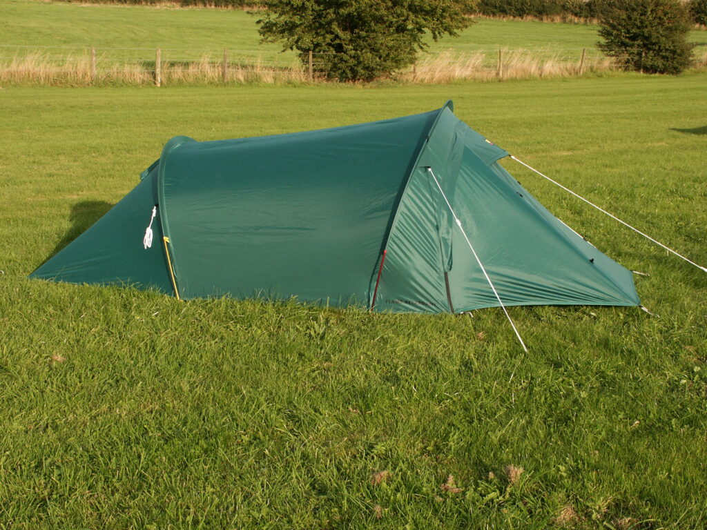 Green tunnel tent staked on a grassy field with tensioned guy lines and a fence and trees along the distant horizon.