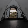 View from inside a grey tent looking out through the open entrance onto a rocky lakeshore with a fallen log, yellow gorse flowers at the water's edge and hills across the calm water.