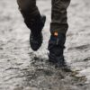 Lower legs wearing brown trousers and black waterproof gaiters with an orange tab and hiking boots splashing through a shallow rocky stream.