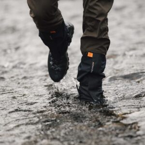 Lower legs wearing brown trousers and black waterproof gaiters with an orange tab and hiking boots splashing through a shallow rocky stream.