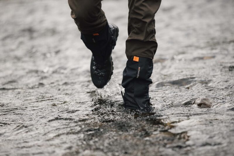 Lower legs wearing brown trousers and black waterproof gaiters with an orange tab and hiking boots splashing through a shallow rocky stream.