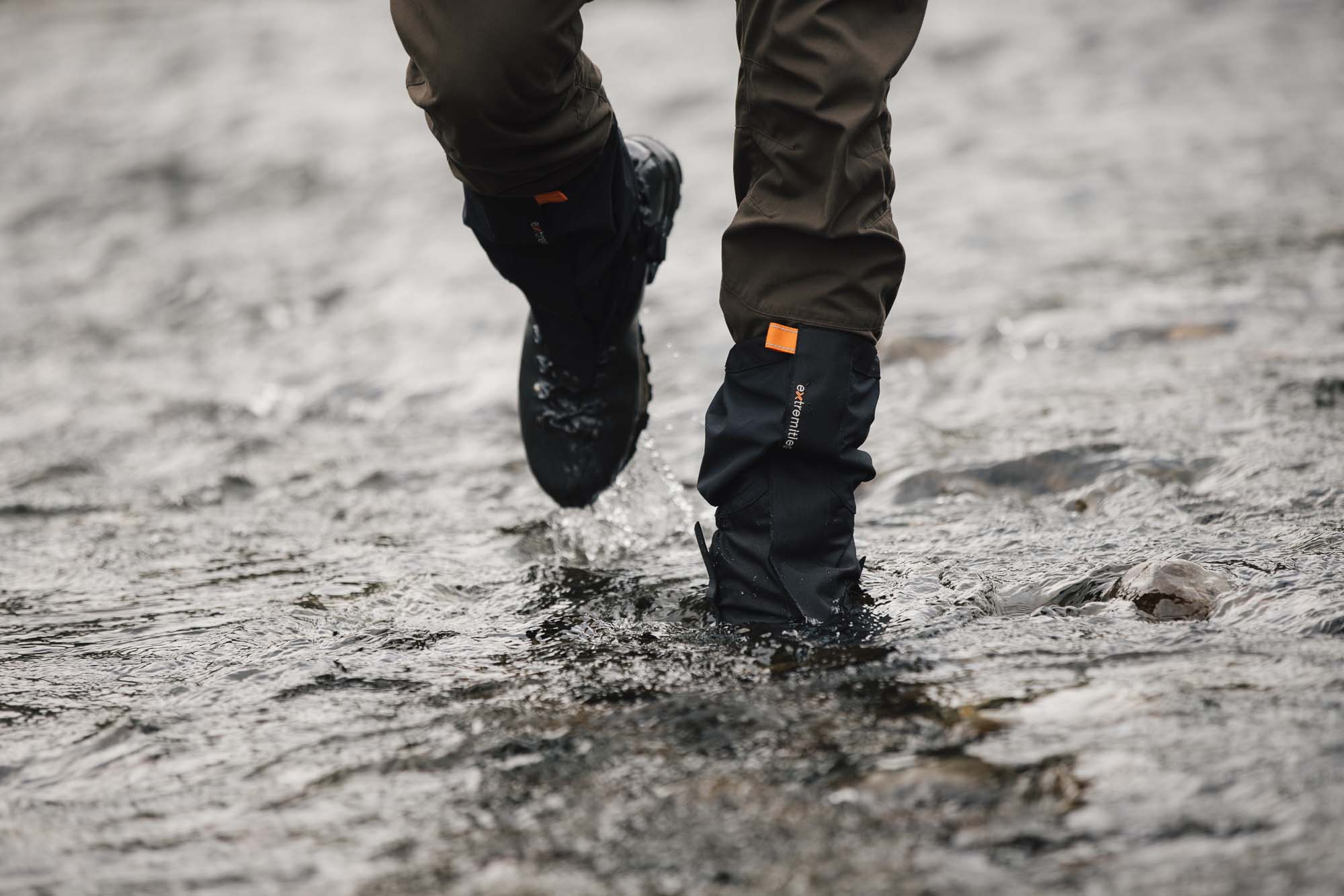 Lower legs wearing brown trousers and black waterproof gaiters with an orange tab and hiking boots splashing through a shallow rocky stream.