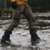 Person's lower body in brown cargo trousers, black gaiters with orange drawcords and hiking boots stepping across a shallow rocky stream with water splashing around the boots