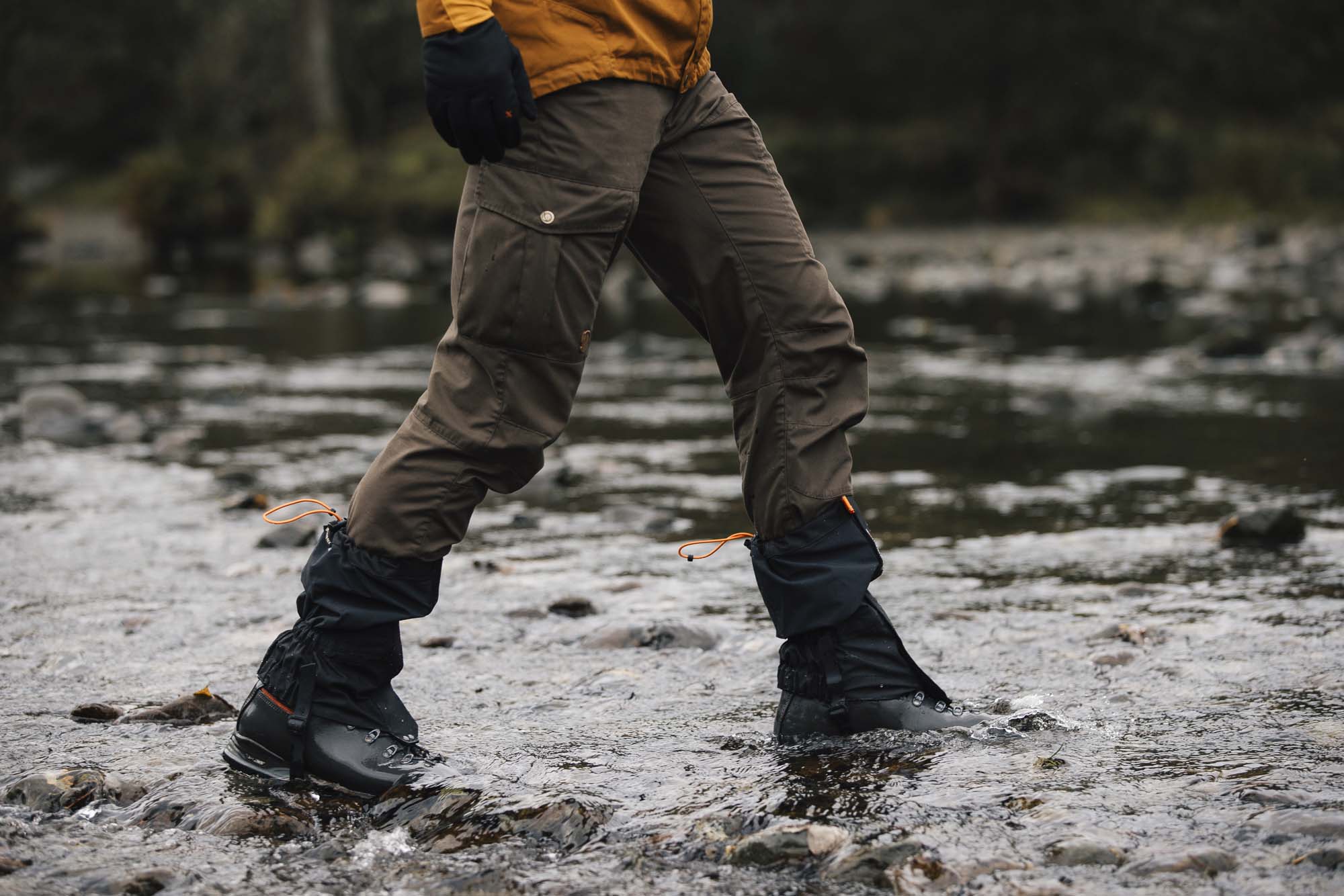 Person's lower body in brown cargo trousers, black gaiters with orange drawcords and hiking boots stepping across a shallow rocky stream with water splashing around the boots