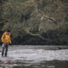 Person in a yellow jacket, black beanie and waders walking through a shallow, pebble-strewn river holding a metal water bottle, with low, mossy trees overhanging the bank in the background.