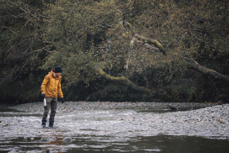 Person in a yellow jacket, black beanie and waders walking through a shallow, pebble-strewn river holding a metal water bottle, with low, mossy trees overhanging the bank in the background.