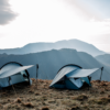 Two blue backpacking tents with extended vestibules held by trekking poles pitched on a grassy mountain plateau, backlit by a bright sky and layered hazy mountain ridges in the distance.