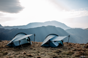 Two blue backpacking tents with extended vestibules held by trekking poles pitched on a grassy mountain plateau, backlit by a bright sky and layered hazy mountain ridges in the distance.