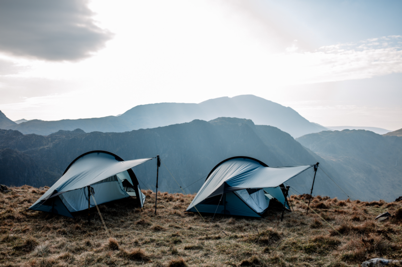 Two blue backpacking tents with extended vestibules held by trekking poles pitched on a grassy mountain plateau, backlit by a bright sky and layered hazy mountain ridges in the distance.