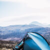 Blue dome tent pitched on windswept grassland with a person at the open door putting on boots, set against rolling heather-covered hills and distant mountains under a pale sky.