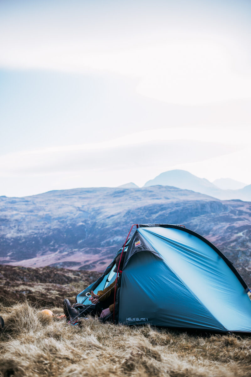 Blue dome tent pitched on windswept grassland with a person at the open door putting on boots, set against rolling heather-covered hills and distant mountains under a pale sky.