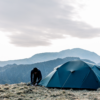 Person crouched securing a dark teal camping tent on a windswept grassy mountain ridge, with layered blue-grey peaks receding into the distance and a large overcast cloud overhead.