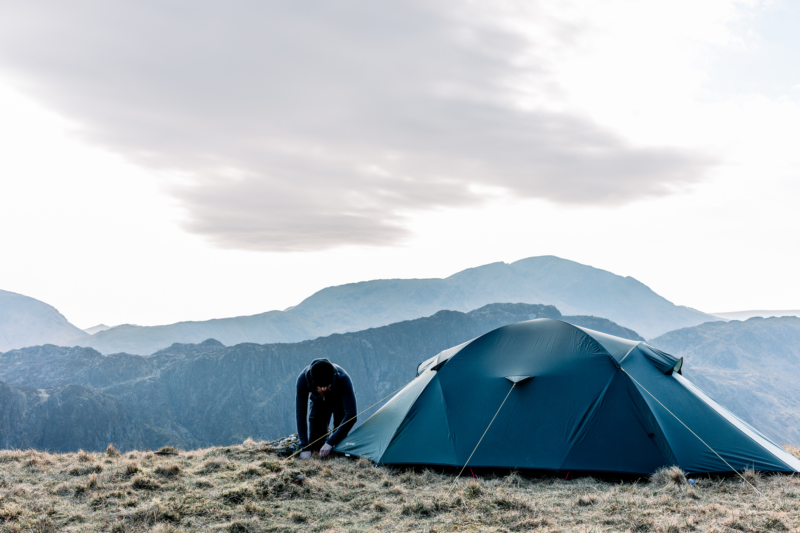 Person crouched securing a dark teal camping tent on a windswept grassy mountain ridge, with layered blue-grey peaks receding into the distance and a large overcast cloud overhead.