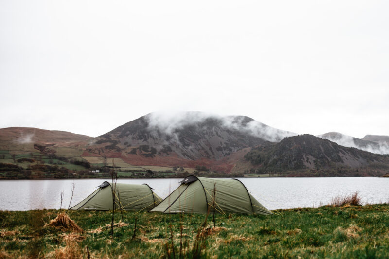 Two olive-green tunnel tents pitched on a grassy lakeshore with calm water and low clouds drifting around rugged hills under an overcast sky