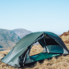 Green two-person dome tent pitched on a dry grassy mountain slope with its front flap open revealing a green sleeping bag inside and distant rocky peaks under a clear blue sky.