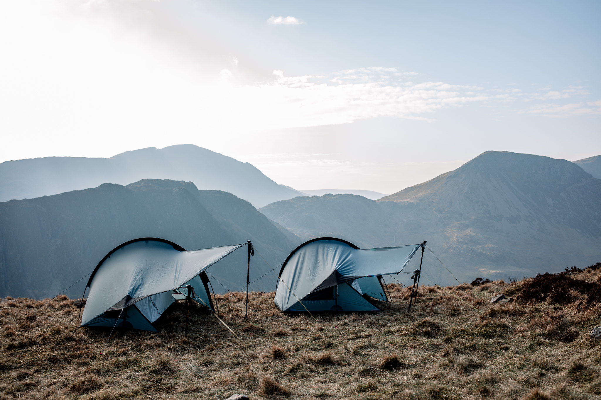 Two low-profile blue-grey tents staked on a windswept grassy ridge, their curved rainflies propped with trekking poles and guylines, with layered misty mountains and a pale morning sky in the background.