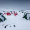 Snow-covered camp of red and green dome tents scattered across a snowy mountain valley, several partially buried in fresh snow with one tent showing a person peering from the entrance.