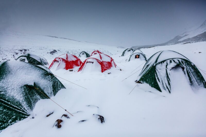 Snow-covered camp of red and green dome tents scattered across a snowy mountain valley, several partially buried in fresh snow with one tent showing a person peering from the entrance.