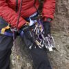 Climber's torso and gloved hands in a red jacket seated on a rock, holding a cluster of silver carabiners, colourful slings and quickdraws clipped to a blue-and-yellow harness with a braided rope running through.