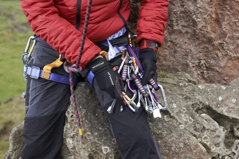 Climber's torso and gloved hands in a red jacket seated on a rock, holding a cluster of silver carabiners, colourful slings and quickdraws clipped to a blue-and-yellow harness with a braided rope running through.