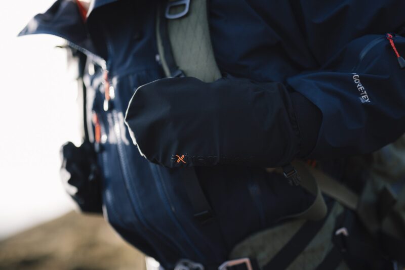 Close-up of a hiker wearing a dark Gore‑Tex jacket with a black insulated mitten bearing an orange 'x' logo resting over a backpack strap