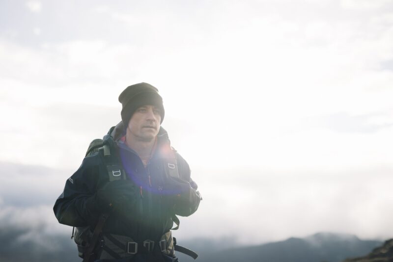 Hiker in a dark beanie and insulated jacket carrying a backpack, standing on a ridge with a bright overcast sky, distant mountains and a faint lens flare across his chest.