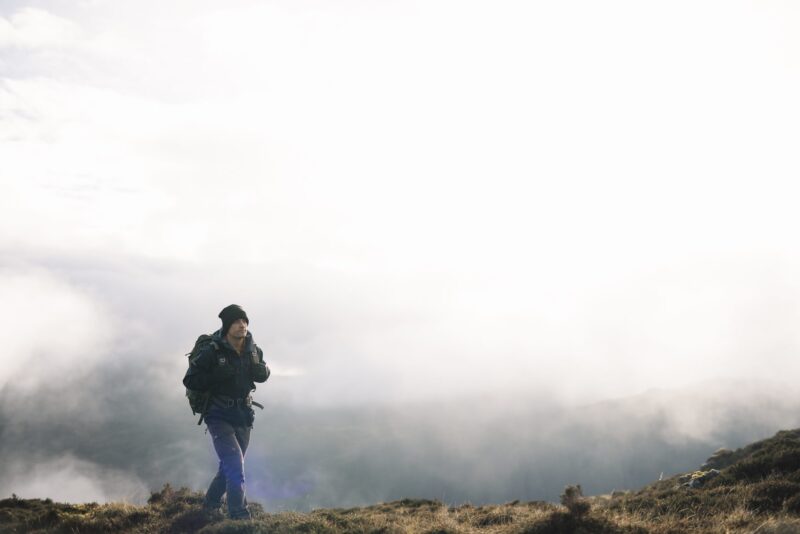 A lone hiker in a black beanie and dark jacket carrying a large backpack walks across grassy, fog‑shrouded mountain terrain under a bright, overcast sky.