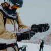 Skier in a beige-and-white jacket wearing a black helmet, yellow goggles and a snow-speckled neck gaiter adjusts grey-and-black gloves next to ski poles on a snowy slope.