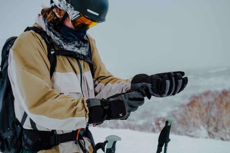 Skier in a beige-and-white jacket wearing a black helmet, yellow goggles and a snow-speckled neck gaiter adjusts grey-and-black gloves next to ski poles on a snowy slope.
