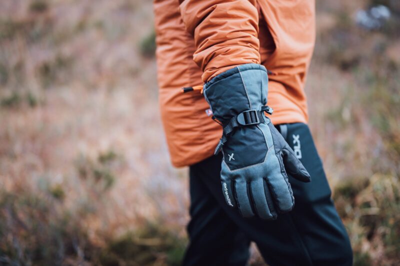 Close-up of a person in an orange jacket showing a grey insulated glove with an adjustable black strap, set against a blurred brown-green outdoor background.