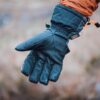 Close-up of an outstretched black insulated winter glove with an orange drawcord and small logo, worn by a person in an orange jacket against a soft-focus outdoor background.