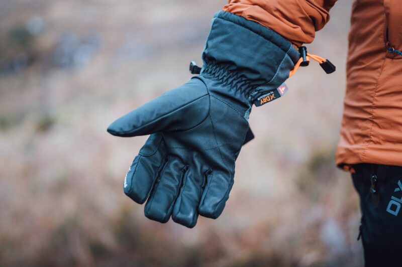 Close-up of an outstretched black insulated winter glove with an orange drawcord and small logo, worn by a person in an orange jacket against a soft-focus outdoor background.