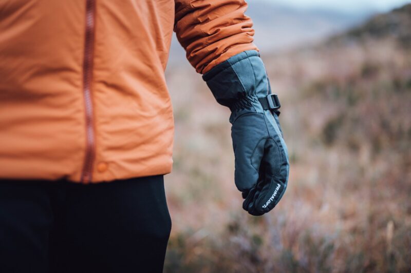 Close-up of a person wearing an orange jacket with their arm at their side in a dark insulated glove with a buckle strap, set against a blurred grassy outdoor background.