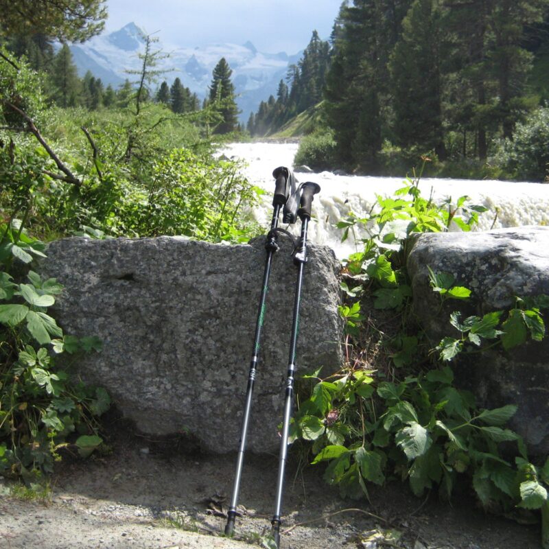 Two trekking poles leaning against a large granite boulder beside a fast-flowing mountain stream, surrounded by green foliage and pine trees with snow-capped peaks visible in the distance.