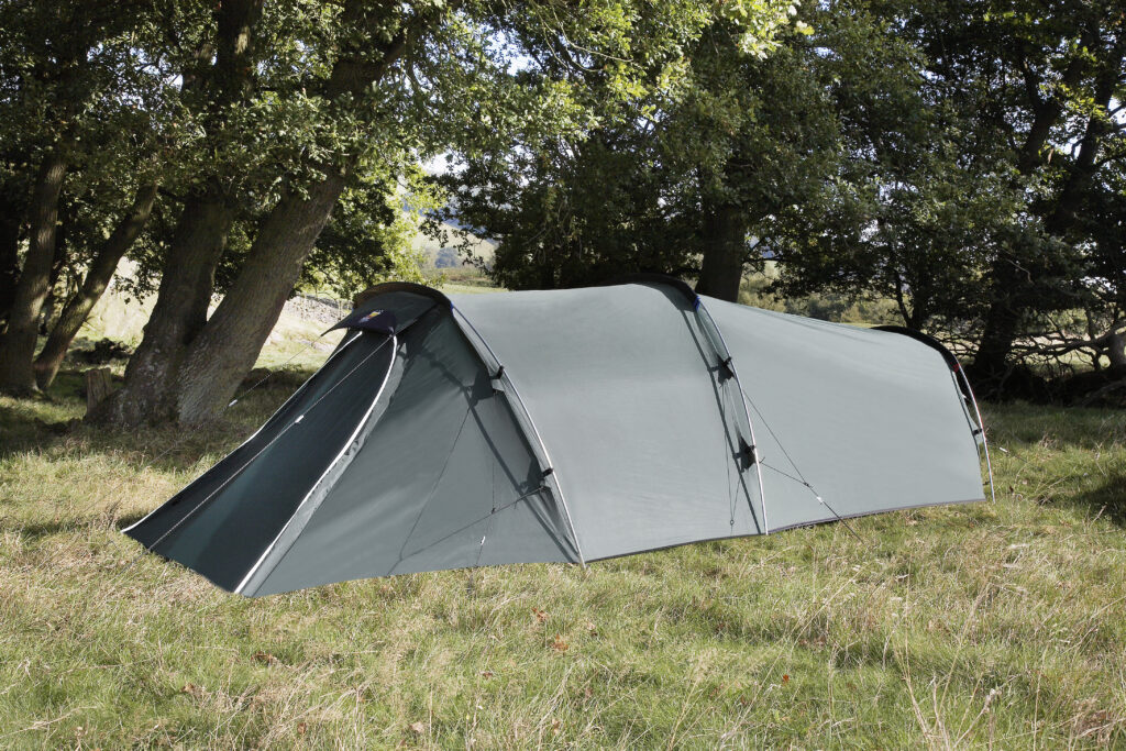 Long grey tunnel-style camping tent with a covered vestibule pitched on a grassy field beneath large deciduous trees