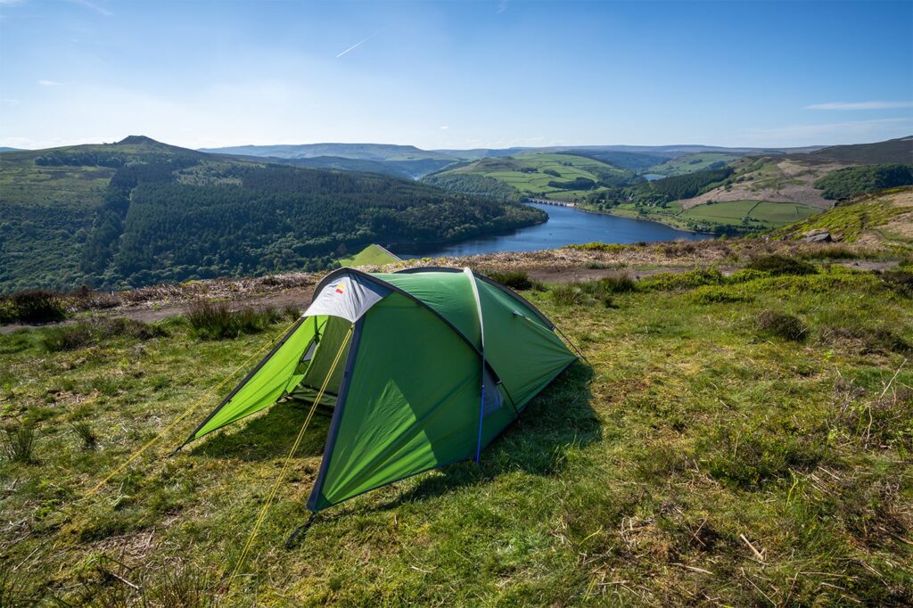 Green backpacking tent pitched on a grassy hilltop overlooking a blue reservoir with a dam and forested, rolling hills under a clear blue sky.