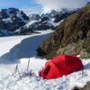 A bright red mountaineering tent pitched in deep snow beside a rocky ridge with skis and poles propped nearby, set against a sweeping glacier and jagged snow-covered mountains under a blue sky.